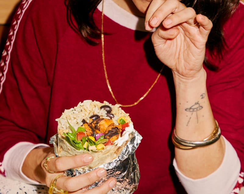 Hands holding a foil-wrapped burrito filled with rice, beans, grilled meat, lettuce and vegetables