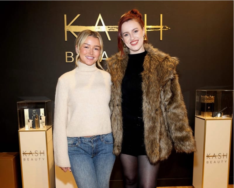 Two women posing at KASH Beauty event with product displays and gold branding