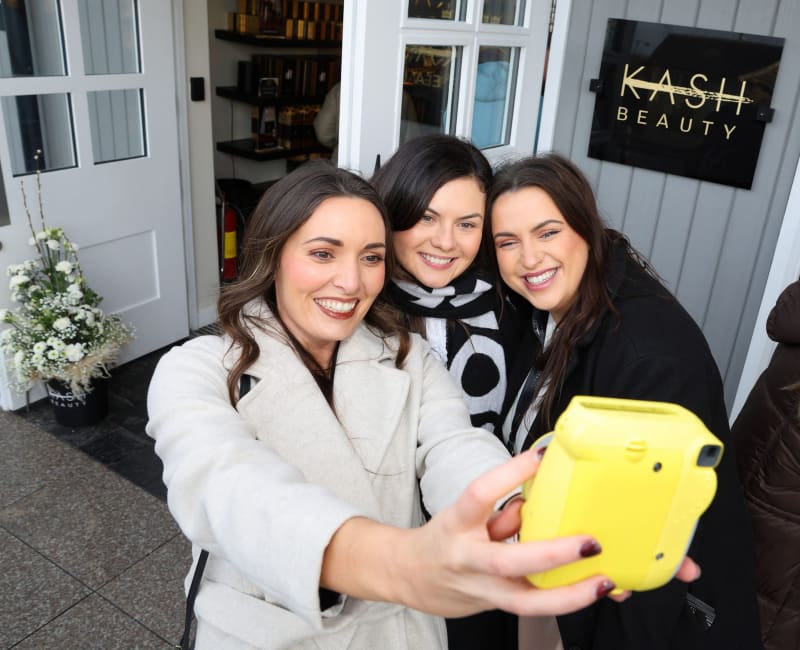 Three smiling women taking a selfie with a yellow instant camera outside KASH Beauty salon