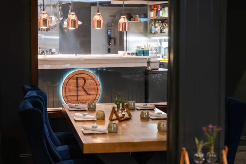 Upscale restaurant interior with wooden dining table, blue velvet chairs, and open kitchen with copper lamps