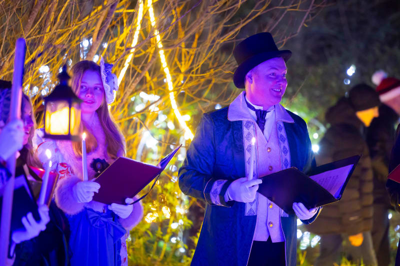 Victorian-dressed carolers singing outdoors at night with lanterns and Christmas lights in background
