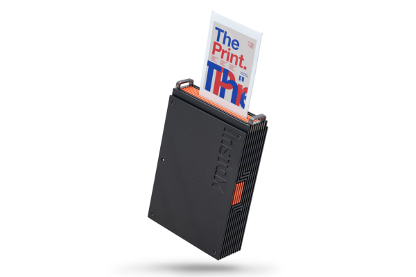 Instax portable photo printer in black with orange accents, printing a page with 'The Print.' text