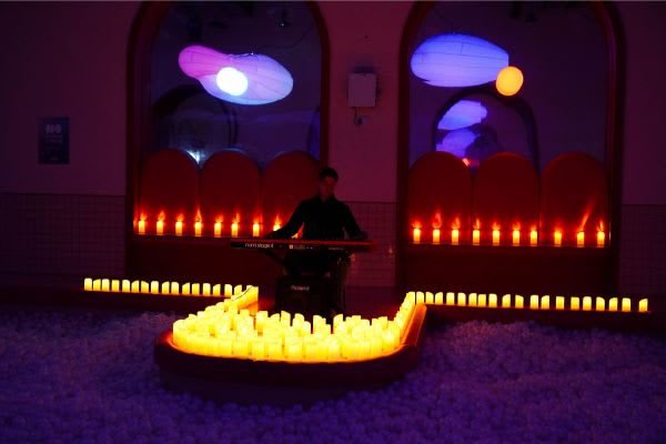 Pianist performing keyboard surrounded by LED candles in atmospheric purple-lit venue with ball pit floor