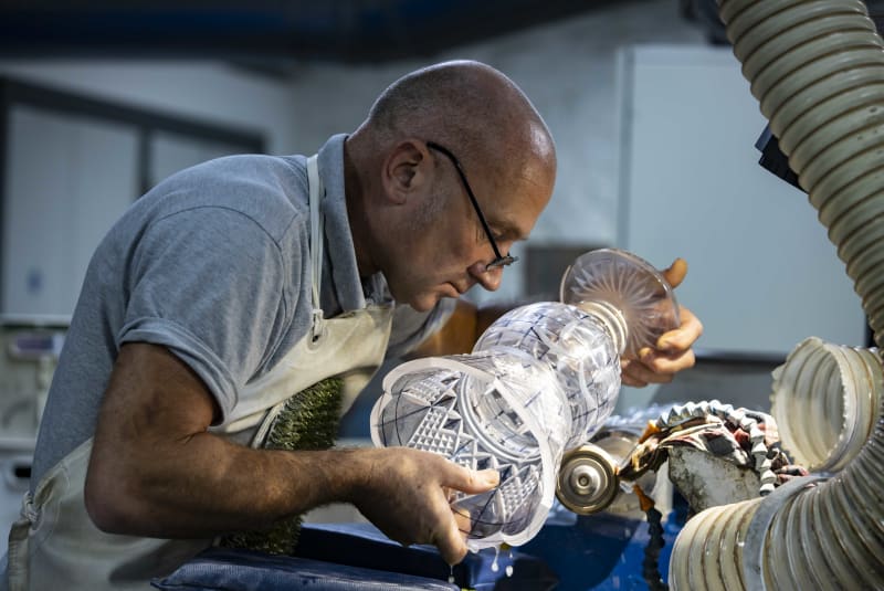 Craftsman cutting intricate geometric patterns into a crystal vase at a grinding wheel in workshop