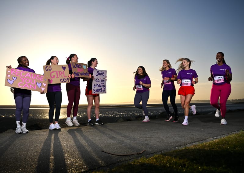 Eight women in purple t-shirts at a running event, four holding motivational signs, four jogging with race bibs