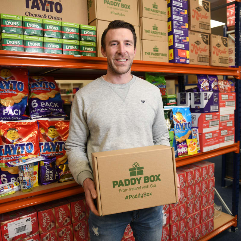 Mark Loftus holding a Paddy Box in warehouse surrounded by Irish snacks including Tayto crisps and Lyons tea