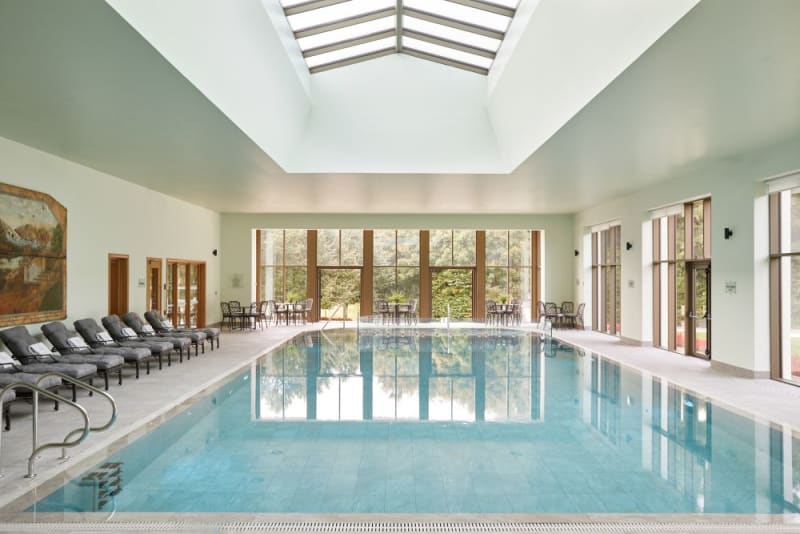 Indoor swimming pool with loungers, large windows overlooking gardens, and skylight in luxury spa setting