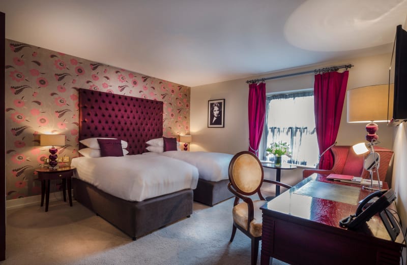 Deluxe hotel room with twin beds, purple tufted headboard, floral wallpaper and magenta curtains