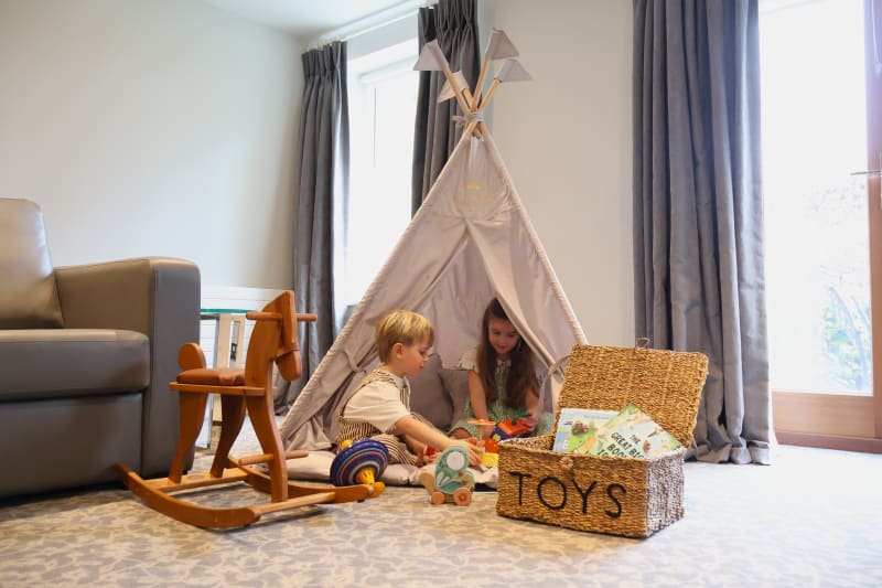 Two children playing with toys near a gray teepee tent in a cozy living room with a wooden rocking horse and toy basket