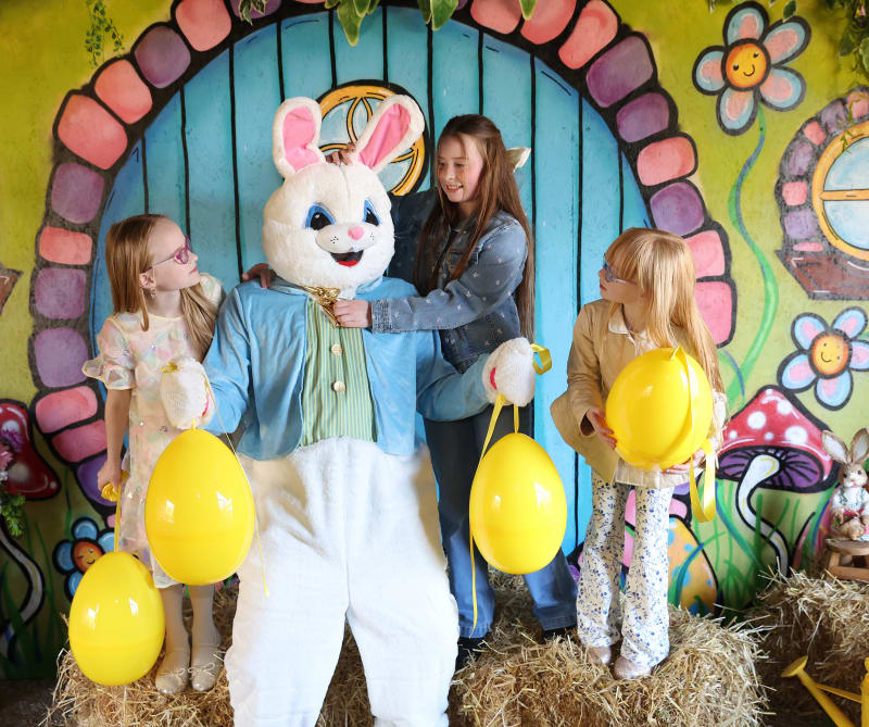 Three girls with yellow egg balloons posing with Easter Bunny character at colorful Easter display