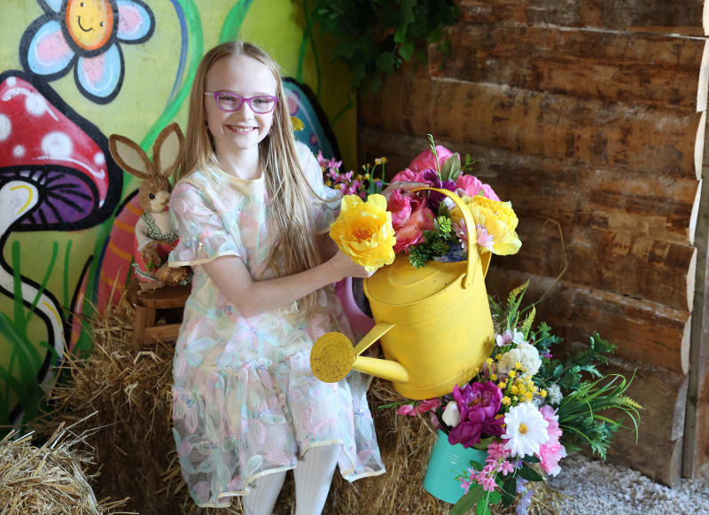 Smiling girl with glasses on hay bales next to yellow watering can filled with colorful flowers at Easter display
