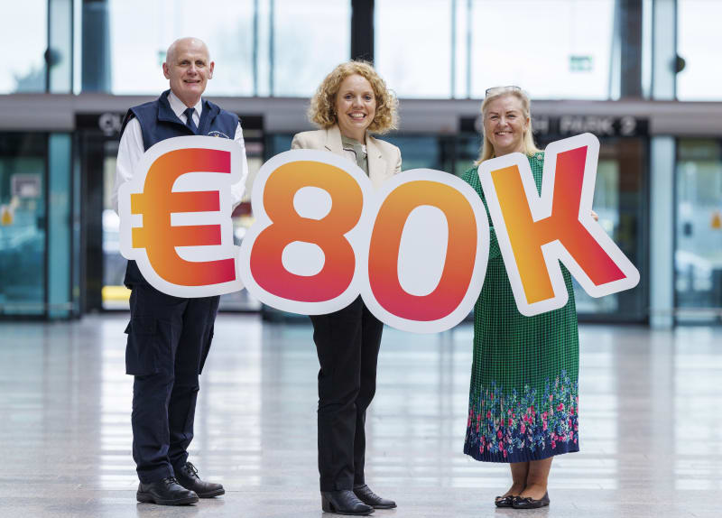 Three people holding a large €80K sign in an airport terminal, celebrating charity fundraising