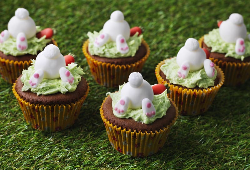 Six chocolate cupcakes with green frosting and white fondant bunnies appearing to dive into the frosting