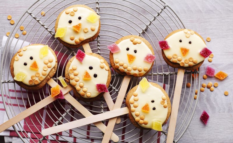 Easter chick cake pops with white icing and candy decorations on wooden sticks arranged on cooling rack
