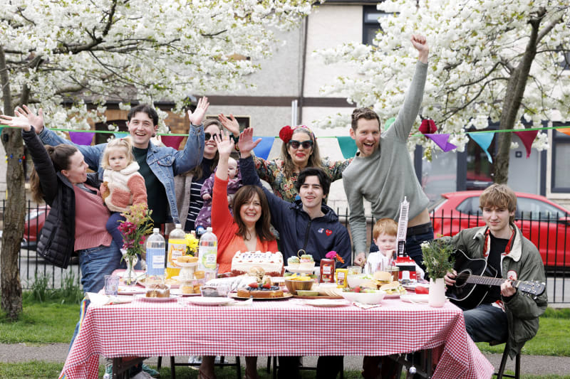 Group of people celebrating at outdoor picnic table with food and drinks, waving at camera under cherry blossom trees