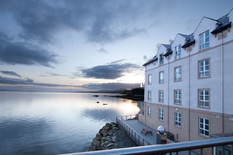Waterfront hotel building at dusk with calm sea, stone embankment, and cloudy sky in background.