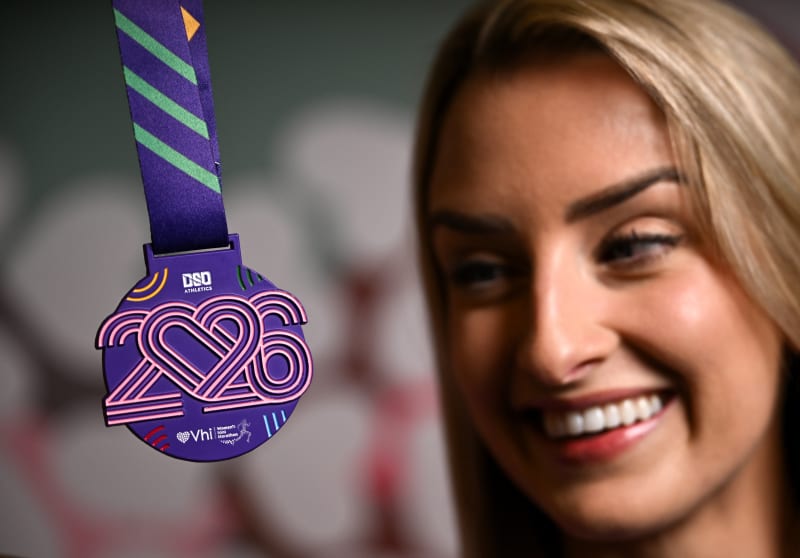 2026 Vhi Women's Mini Marathon finisher medal in purple with pink neon-style numerals, held up by a smiling woman.