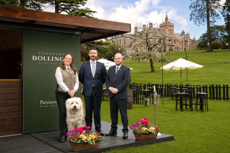 Three hospitality staff and a white fluffy dog pose before a Champagne Bollinger banner at a grand castle estate.