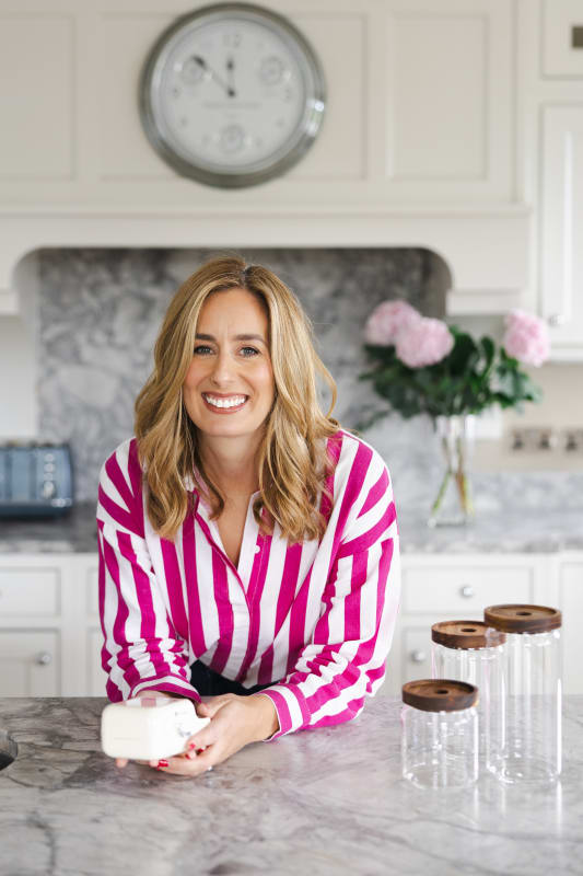 Hazel Burton smiling in a pink striped shirt, leaning on a marble kitchen counter holding a small white product.