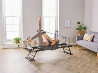 Person using a Pilates reformer machine at home, legs raised, using resistance straps in a bright living room.