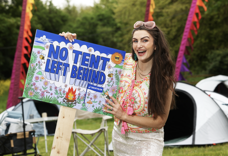 Woman holding a colourful 'No Tent Left Behind' Decathlon campaign sign at a festival campsite.
