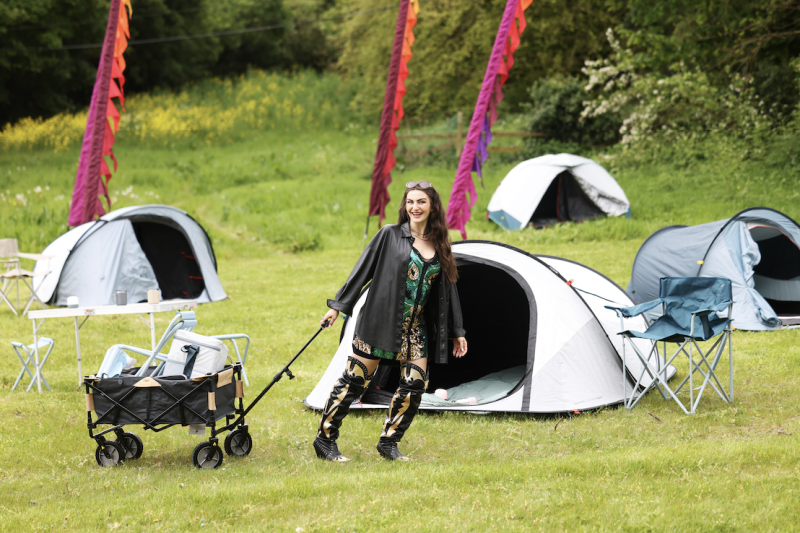 Woman in festival outfit pulling a folding utility wagon at a campsite with white dome tents and colourful streamers.