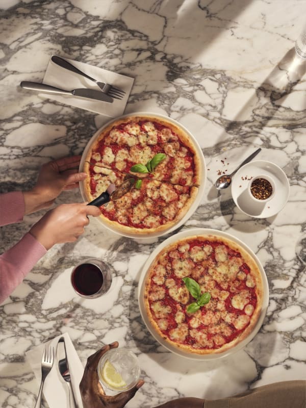 Two Margherita pizzas on a marble table, one being sliced, with red wine, water, and chilli flakes nearby.
