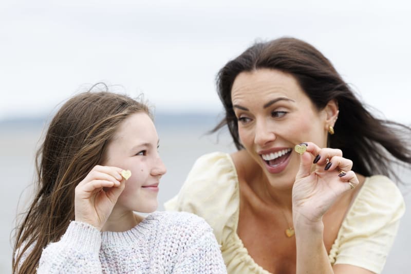 A woman and young girl each hold up a small gold heart-shaped pendant on a windy beach, smiling.