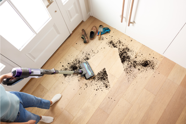 Person using a cordless stick vacuum to clean a large dirt spill on a light wood hallway floor.