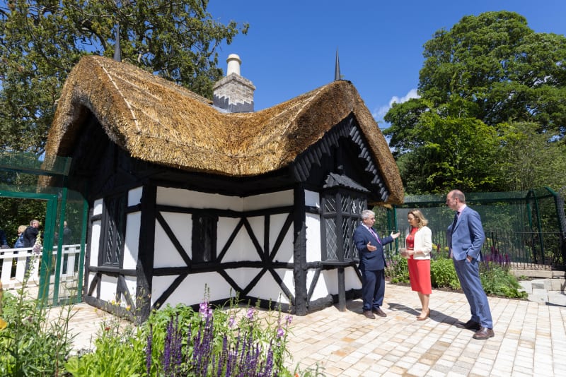 Three people stand beside a restored Tudor thatched cottage with half-timbered walls at Dublin Zoo.