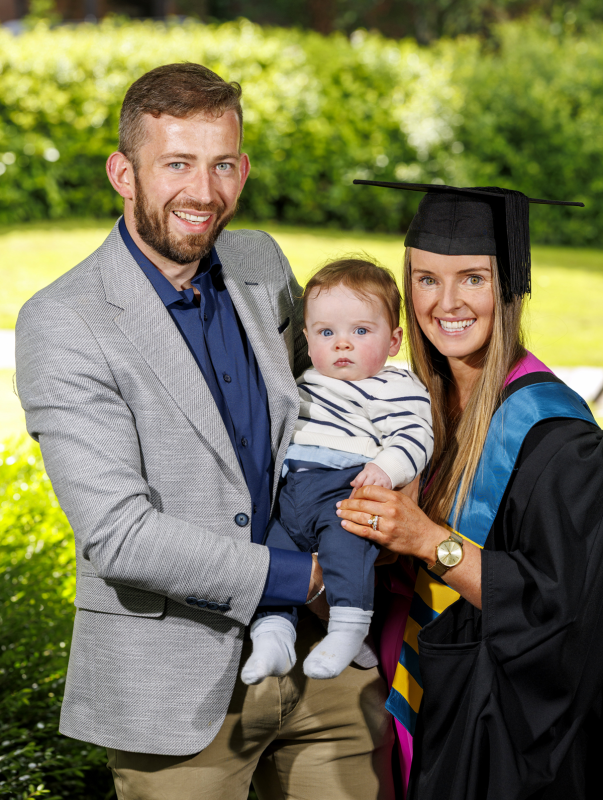 Naomi Carroll in graduation gown and cap with a man in grey blazer and a baby, outdoors in sunny garden.