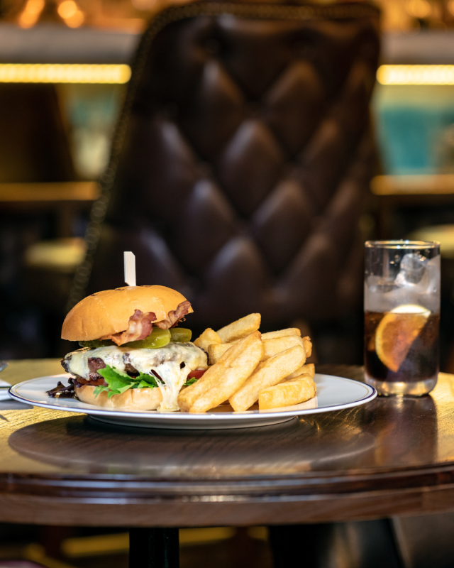 Loaded cheeseburger with bacon and fries on a white plate beside an iced cola drink in a stylish restaurant.