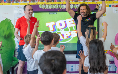 Two men lead an energetic exercise session called Toe Taps with children in a primary school classroom.