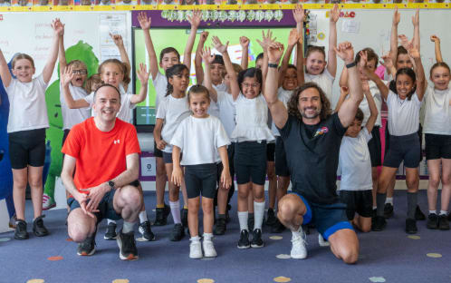 Two men in sportswear kneel in front of a cheering group of primary school children in a classroom.
