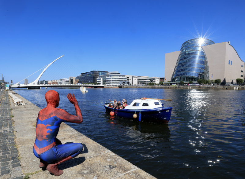 Spider-Man costume character kneels on Dublin's River Liffey quay waving at a passing boat, Samuel Beckett Bridge behind.