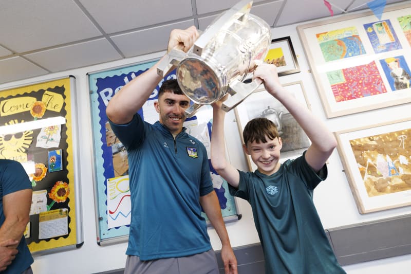 Adult man and young boy smiling as they lift a large crystal GAA trophy together in a school classroom.