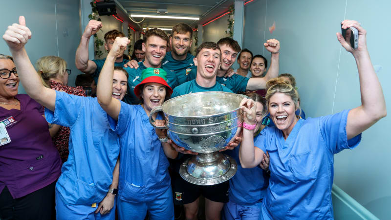 GAA players in green jerseys celebrate with hospital staff holding the Sam Maguire Cup in a hospital corridor.