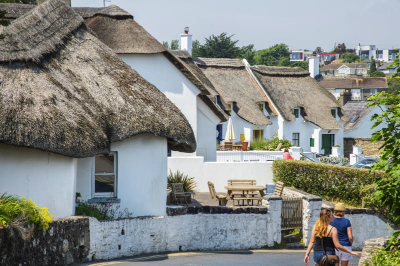 Traditional whitewashed thatched cottages lining a sunny street in Dunmore East, Ireland, with two women walking past.