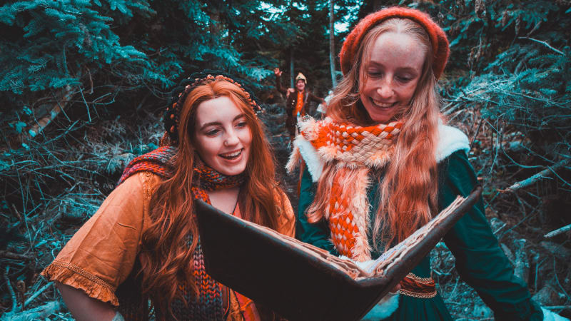 Eabha and Fiadh in medieval fantasy costumes reading an old leather-bound book together in a forest.