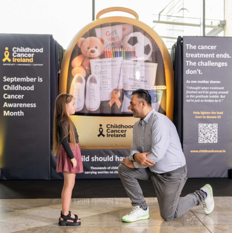 Man kneels to talk with young girl in front of Childhood Cancer Ireland awareness backpack display at Dundrum Town Centre.