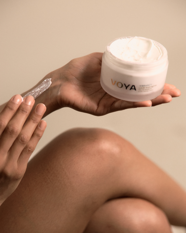 Hand holding open VOYA Vibrancy Luxury Body Butter jar while applying product to skin.