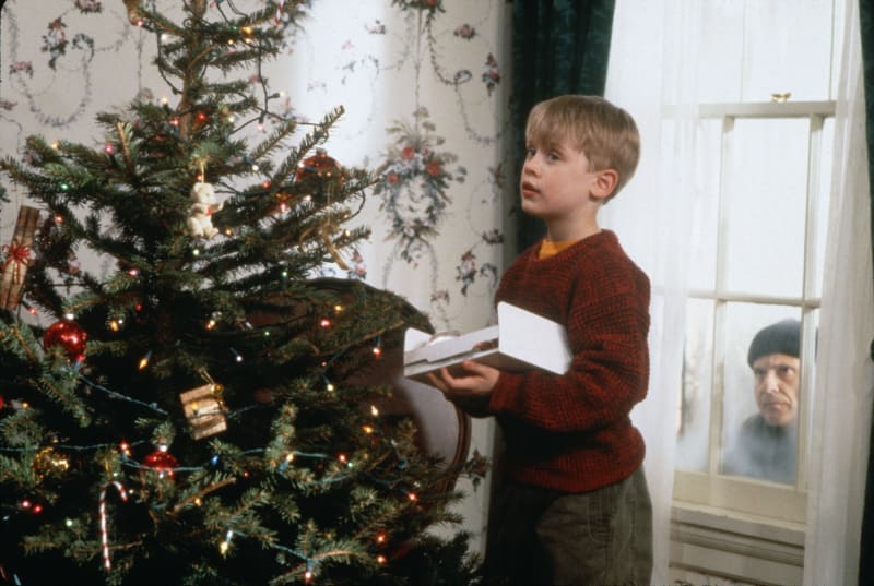 Home Alone: boy in red sweater holds gift box by Christmas tree as man peers through window outside.