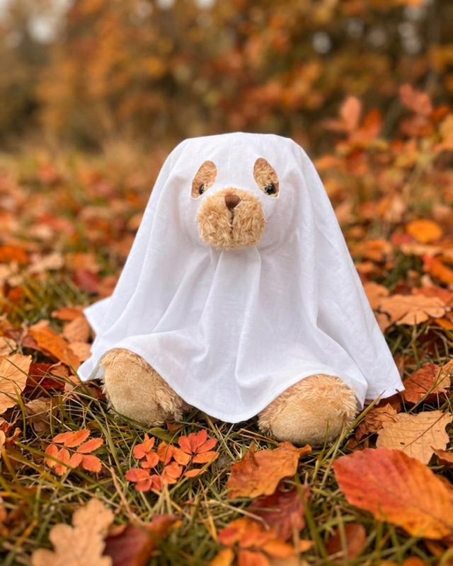 Teddy bear wearing a white ghost Halloween costume sitting among autumn leaves outdoors.