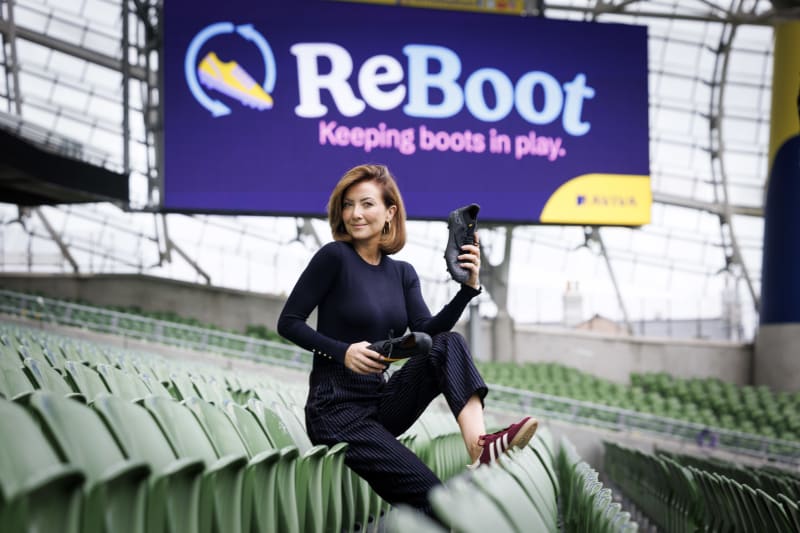 Jennifer Zamparelli holds black football boots in Aviva Stadium with ReBoot campaign branding visible behind her.
