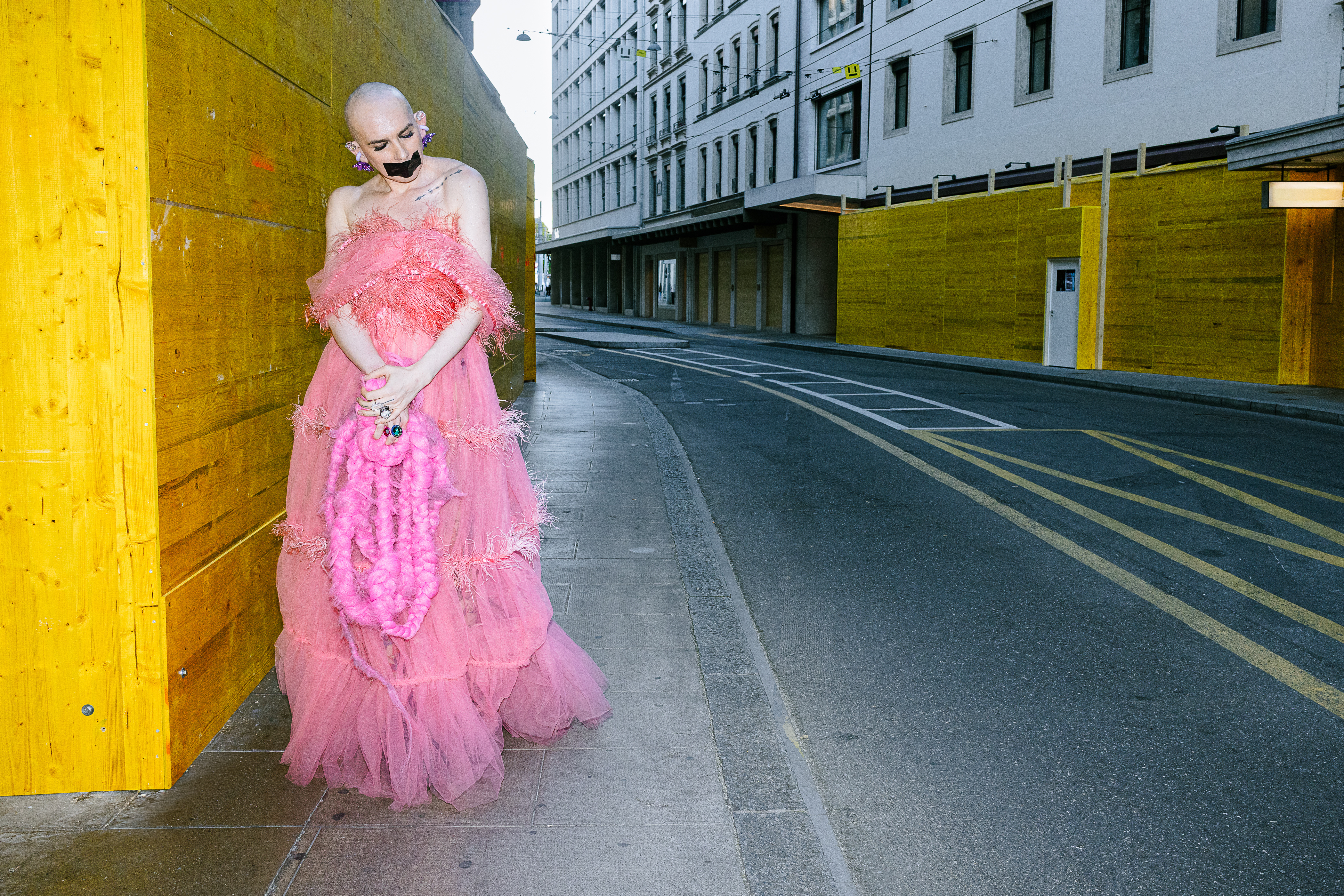 Boginya Von Art wearing a pink dress beside a yellow wall on an urban street.