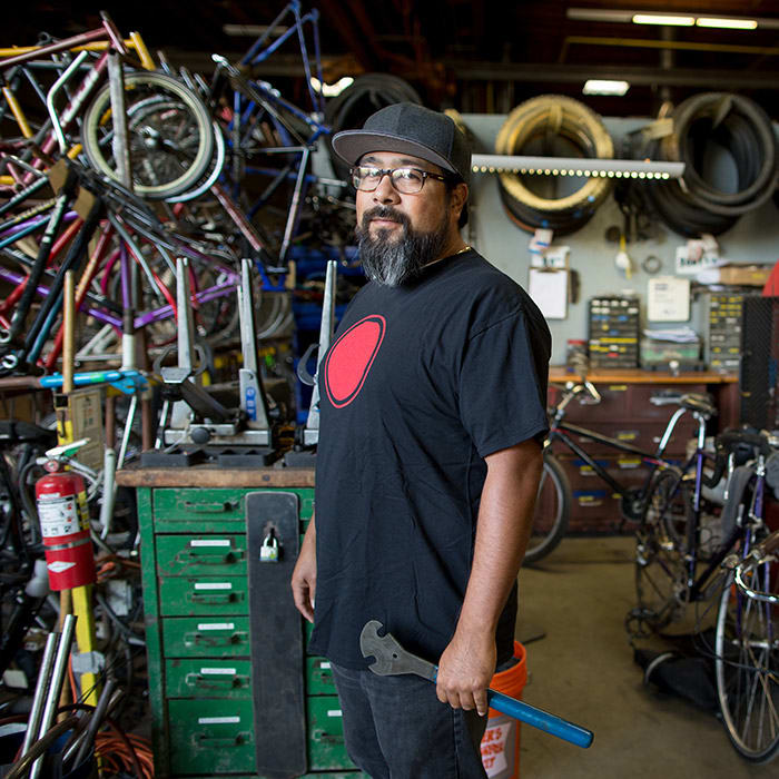 Man standing in his bike repair shop, looking at the camera, with lots of bikes and equipment around him