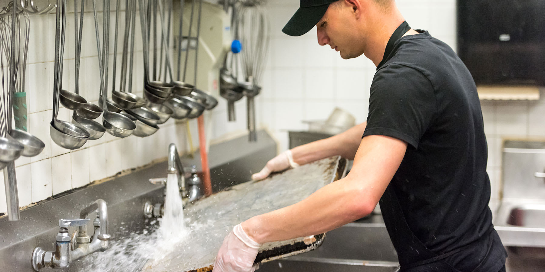 Man washing large baking sheet in the sink of a commercial kitchen.