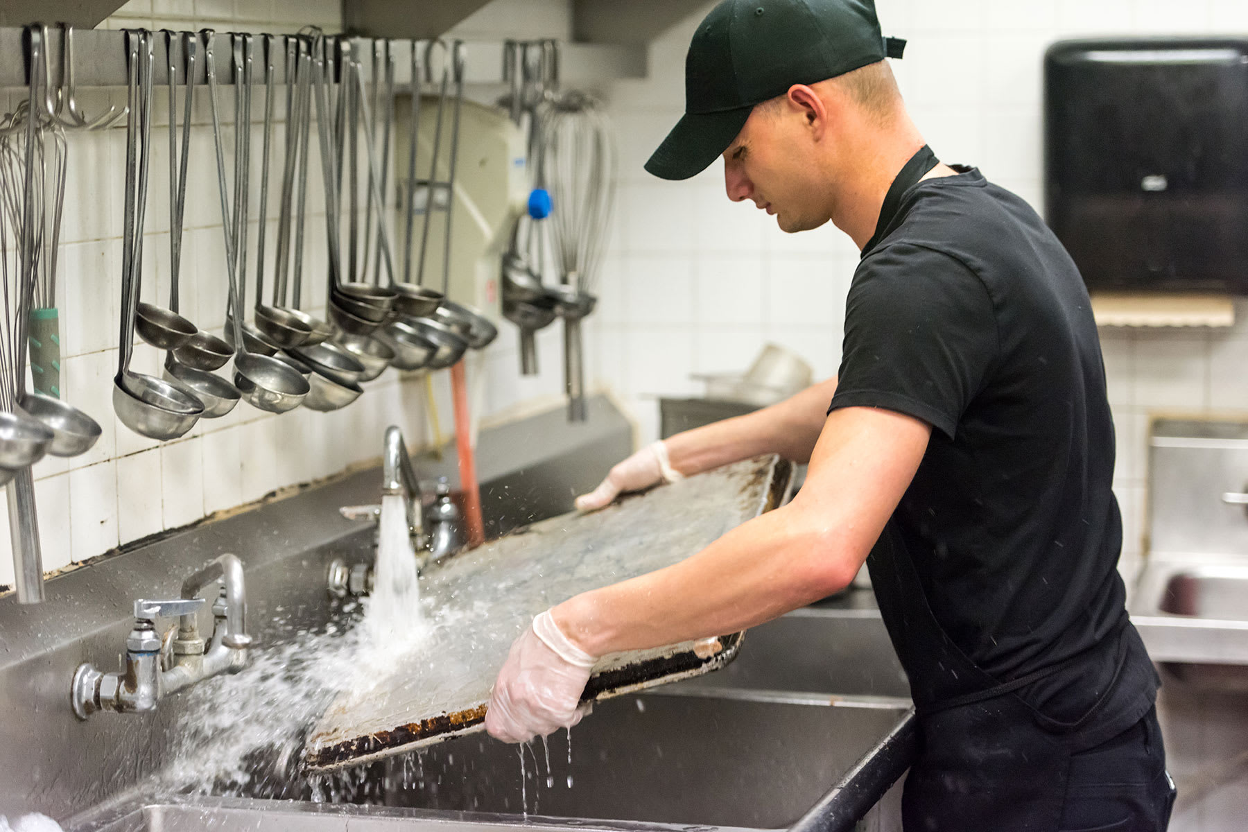 Man washing large baking sheet under the sink in a commercial kitchen.