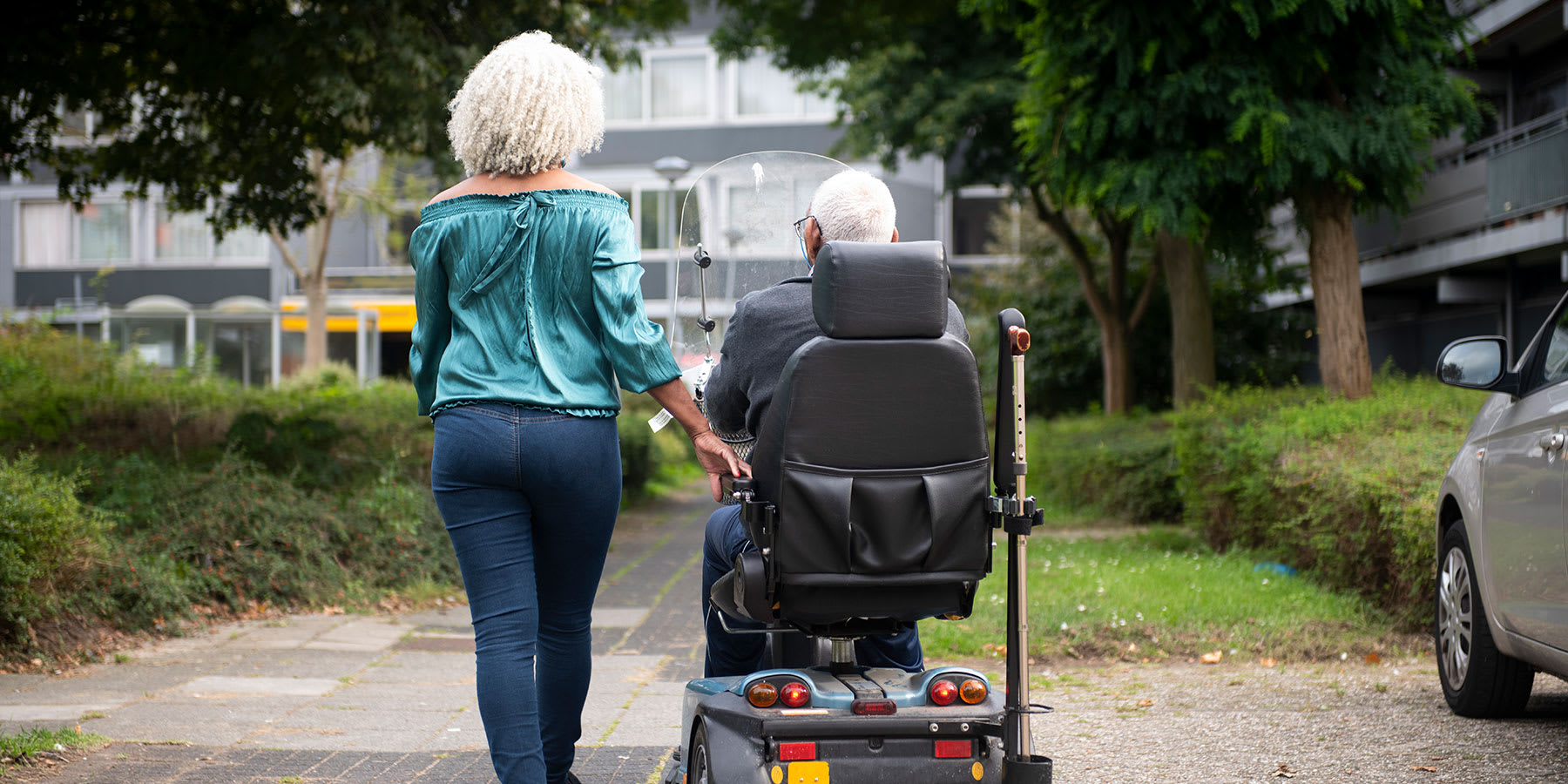 Elder woman walking next to her husband in a motorized wheelchair.