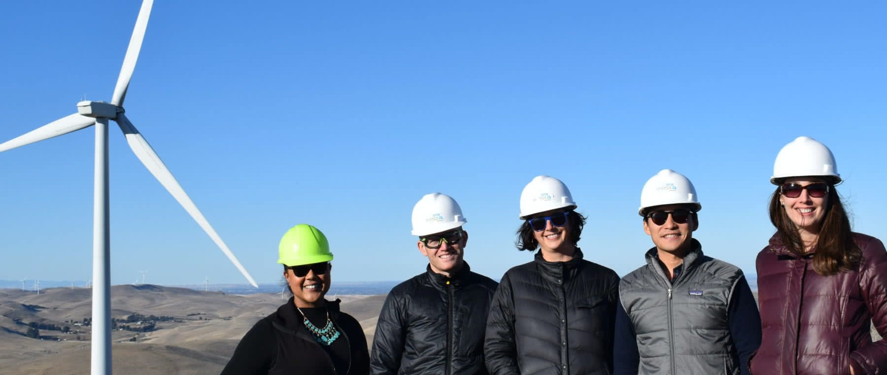 Five employees from the the EBCE team posing in front of a wind turbine with hard hats and sunglasses on.